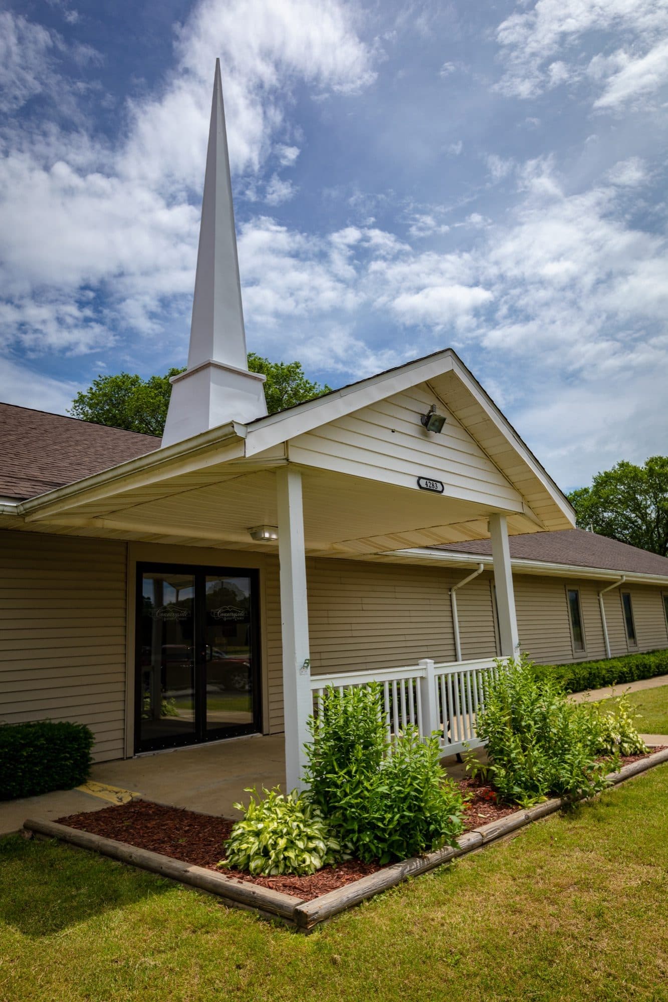 The covered main entrance at Countryside Baptist Church, with the white steeple rising above the porch roof, glass double doors, and planted beds either side of the walk.