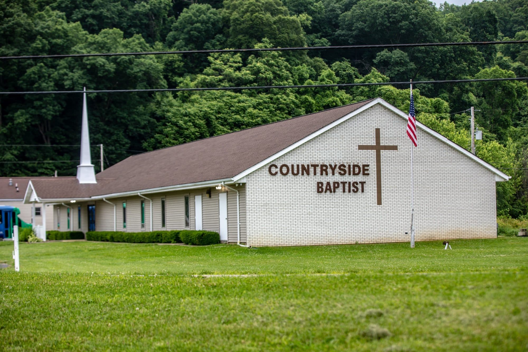 Countryside Baptist Church from the road on a summer afternoon: a white brick gable carrying a tall cross and the words COUNTRYSIDE BAPTIST, a white steeple over the entrance wing, and a wooded hillside rising behind the building.