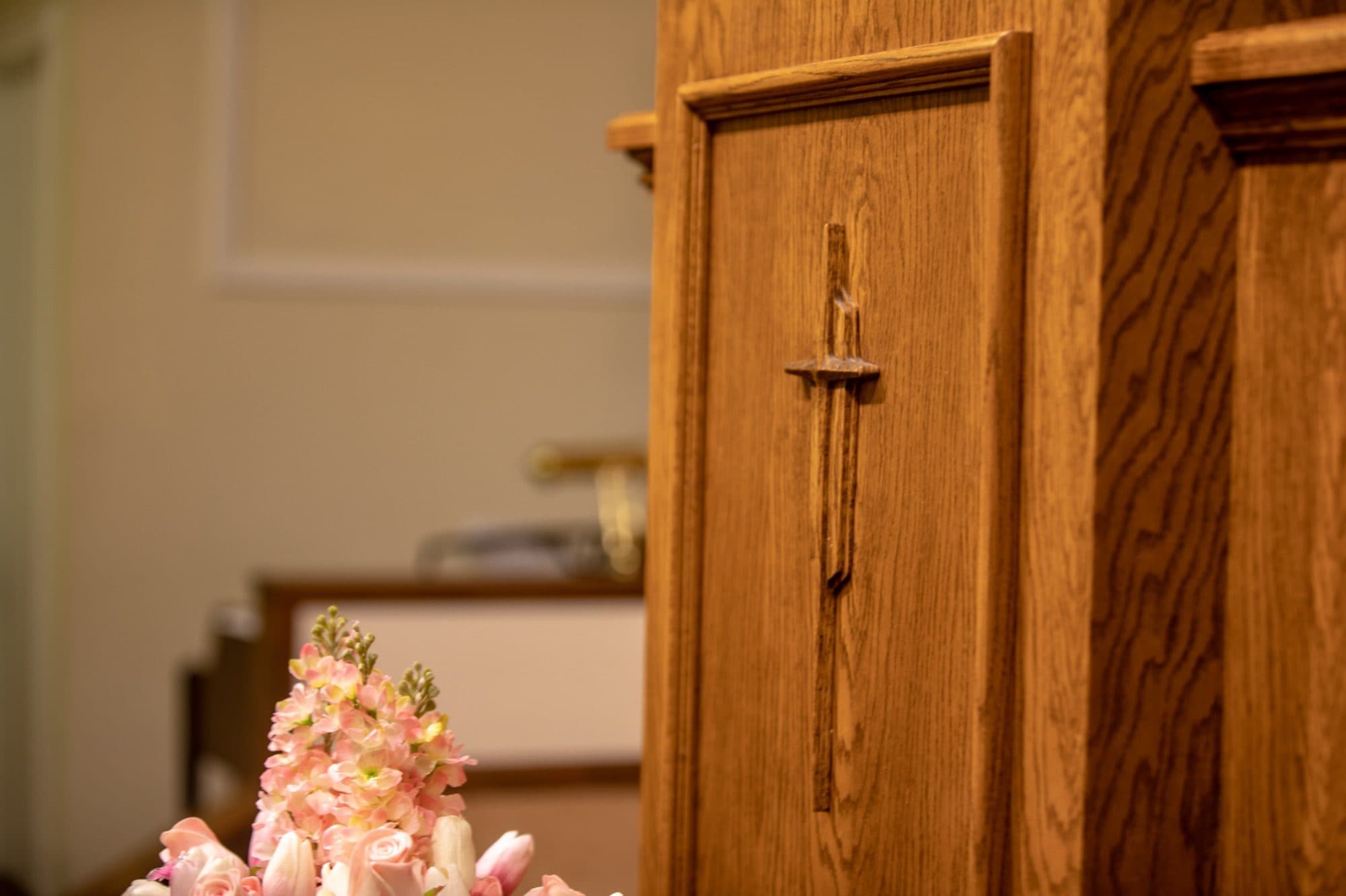 A cross carved into the honey-oak panelling of the pulpit at Countryside Baptist Church, with an arrangement of pink flowers on the communion table in front of it.