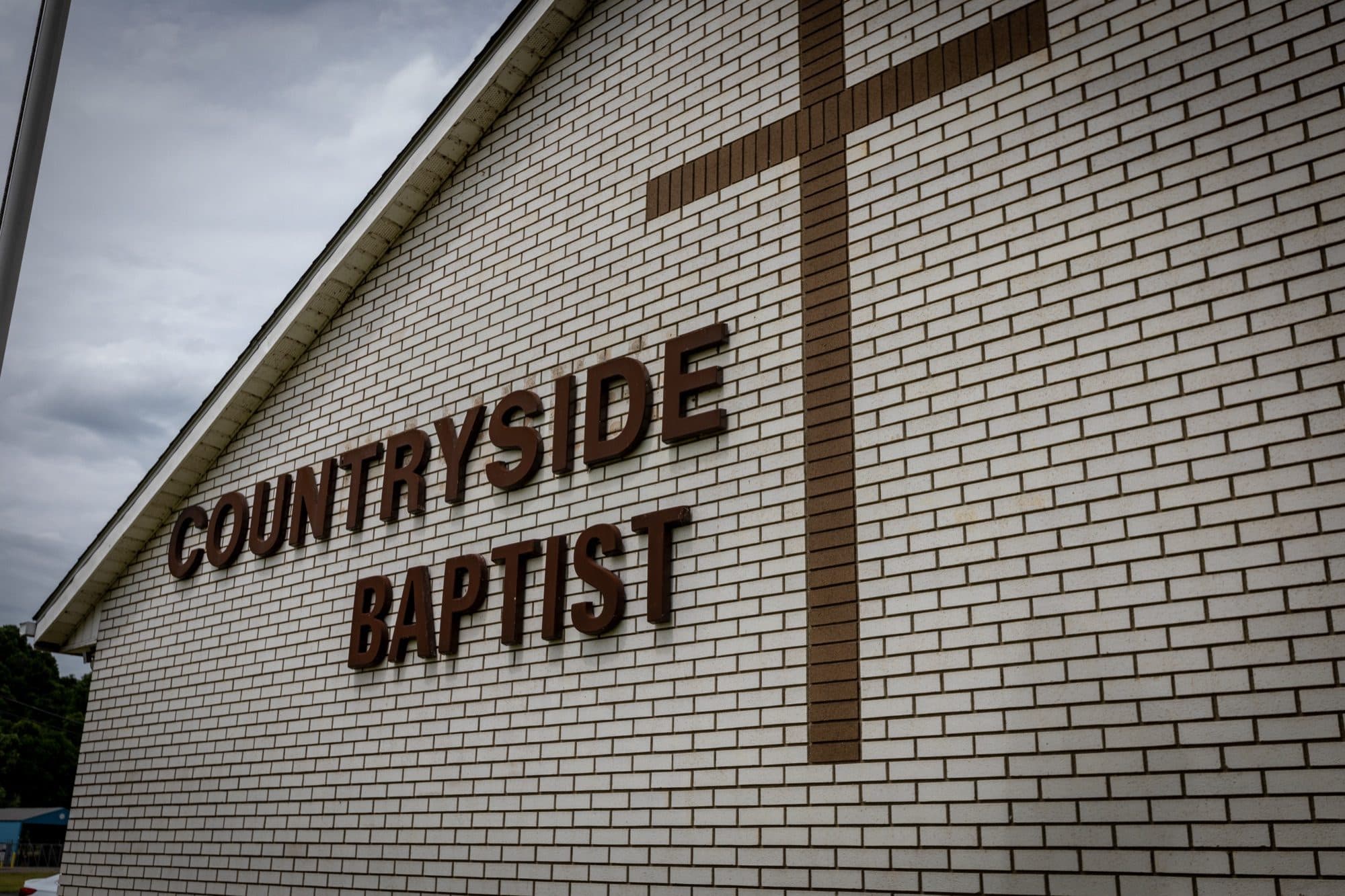 A low view along the brick gable wall of Countryside Baptist Church, with the raised lettering COUNTRYSIDE BAPTIST and a cross laid into the brickwork above it.